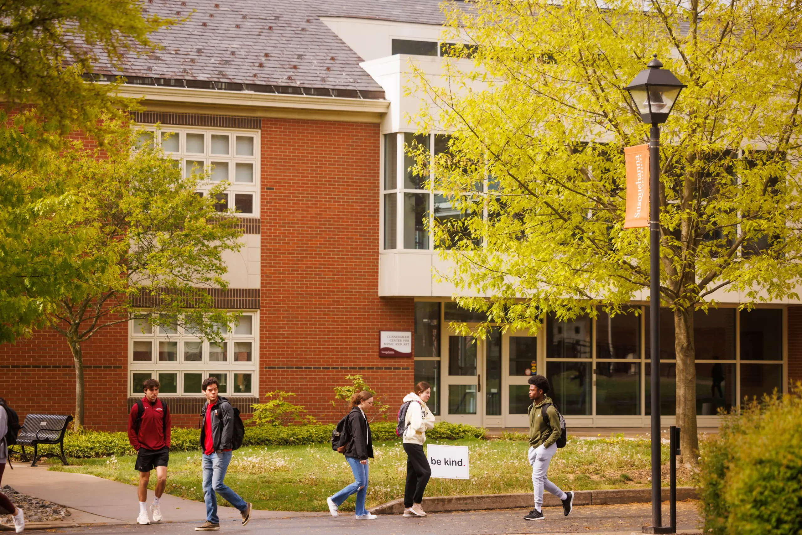 Students walking on a college campus path, surrounded by green trees. A brick building with many windows is in the background. One student holds a white sign with the words