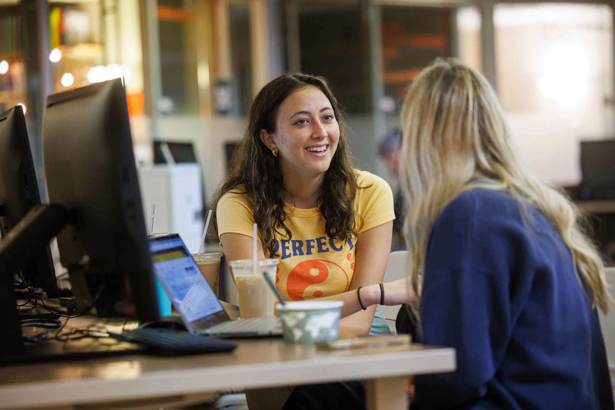 Two people sitting at a table with laptops, smiling and talking. One person is wearing a yellow shirt with lettering, the other a blue sweatshirt. They are in a well-lit room with blurred background. Drinks are on the table.