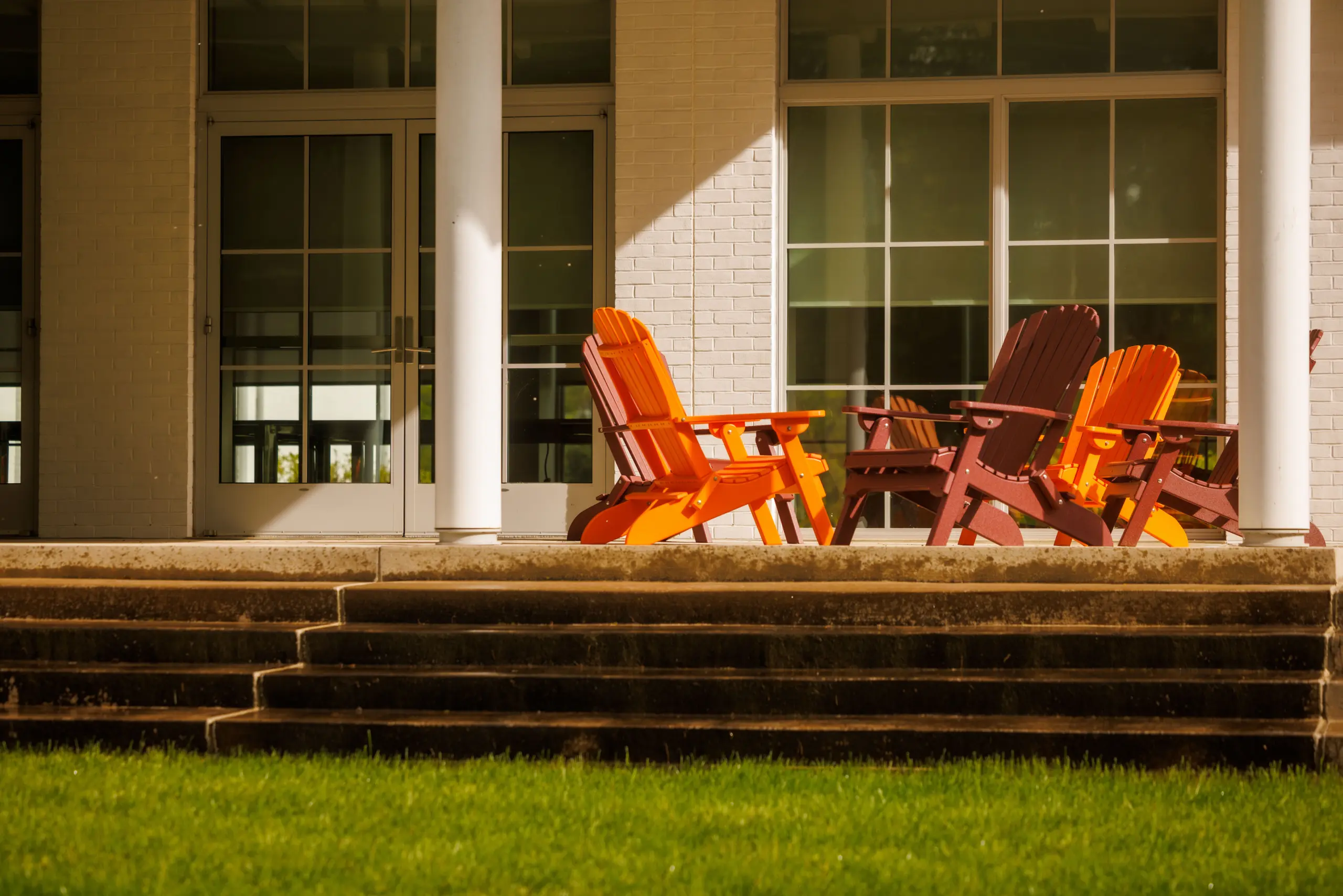 A set of vibrant orange and red Adirondack chairs sit arranged on the patio of a building. The scene is sunlit, with shadows cast on the brick wall and glass doors behind them. Green grass is visible in the foreground.