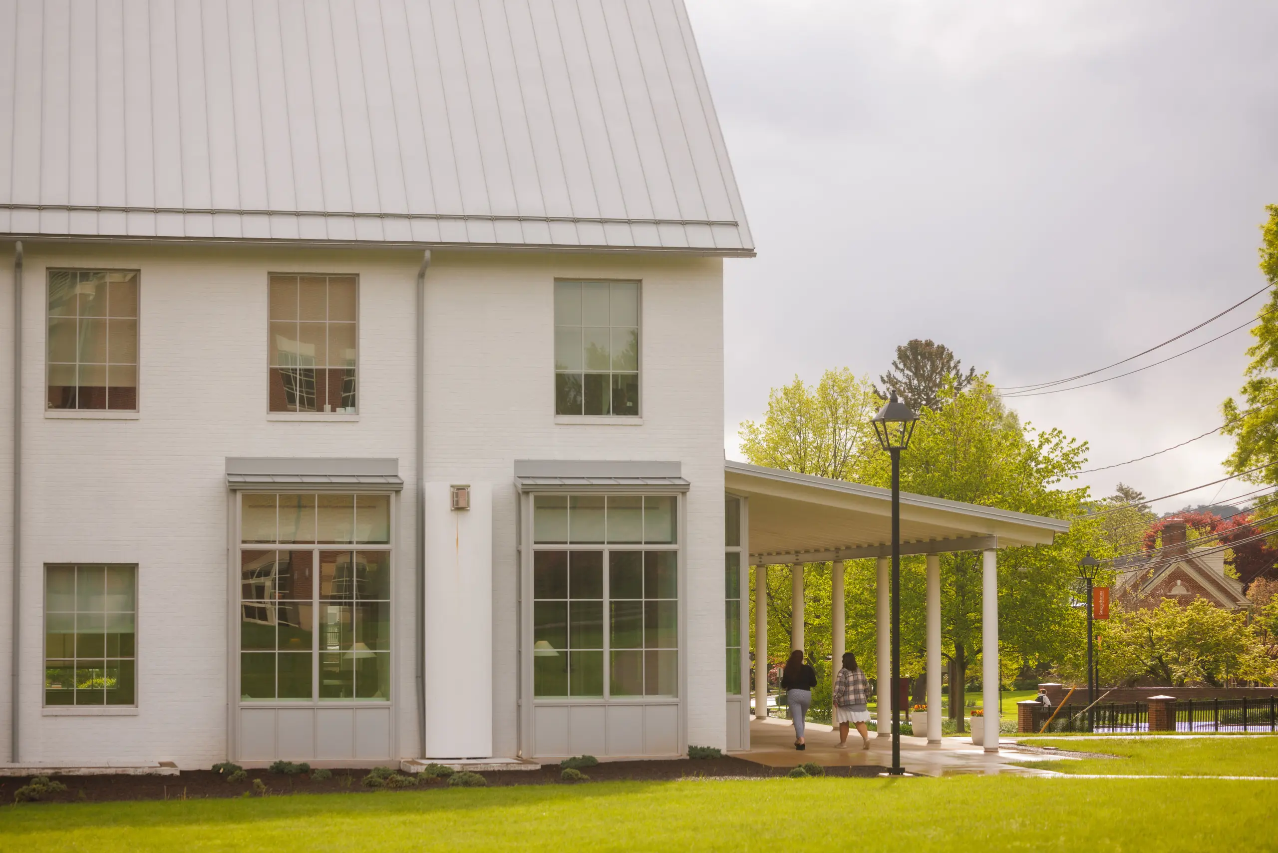 A modern white brick building with large windows and a metal roof is partially visible. Adjacent is a covered walkway. Two people are walking along the path. The background shows trees and a partly cloudy sky.