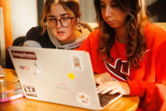 Two young women focus intently on a laptop. One wears glasses and a grey hoodie, while the other wears a red sweatshirt. The laptop is decorated with various stickers, and they appear to be studying or working together at a wooden table.