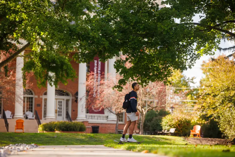 A student walks on a sunny campus path surrounded by greenery and trees. A red brick building with white columns is in the background. The student carries a backpack, and colorful chairs are visible on the grass near the building.