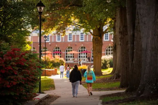 People walk along a tree-lined path on a college campus, leading to a brick building with large arched windows. The scene is bright and sunny, with green grass and foliage surrounding the walkway.