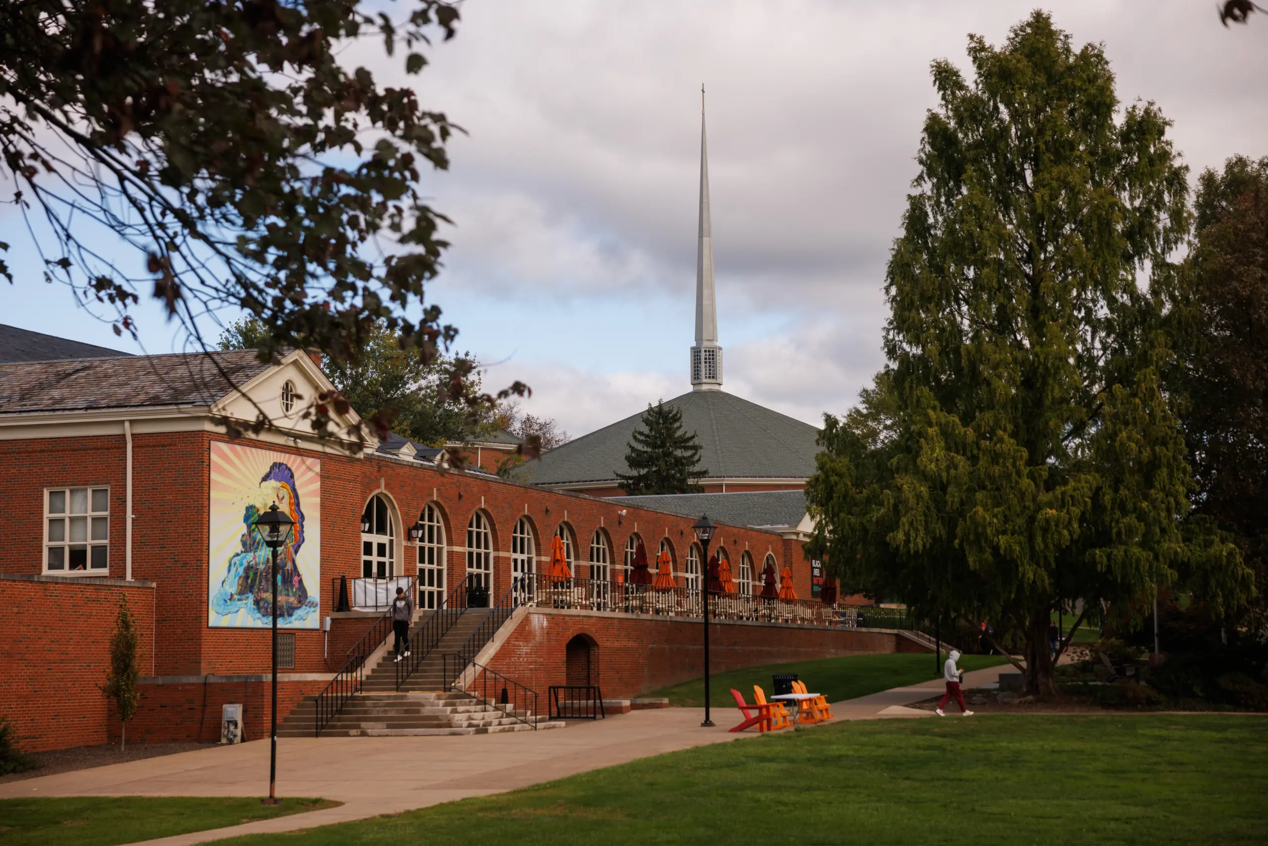 A scenic view of a campus with a red brick building featuring arched windows and a colorful mural. A tall spire is visible in the background. Trees and green grass surround the area, with a few chairs and people walking along pathways.