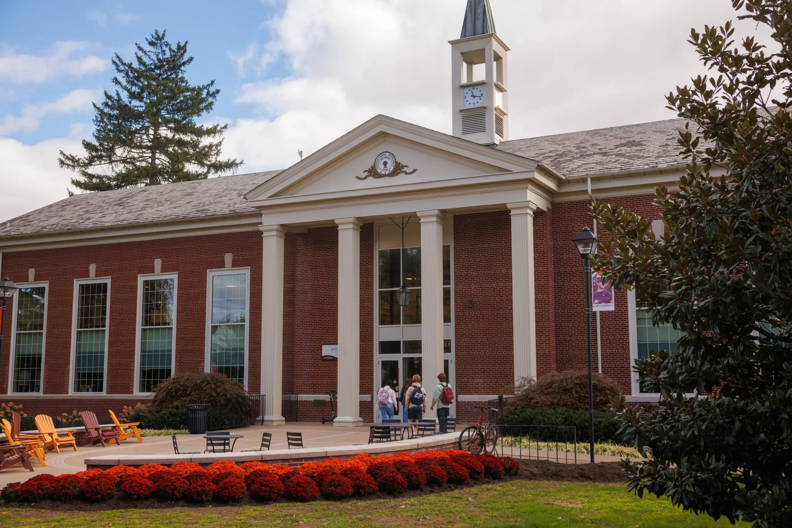 A red-brick building with a clock tower and columns at the entrance. Several people stand in front, and there are Adirondack chairs and tables on the patio. A circular bed of red flowers is in the foreground, under a partly cloudy sky.