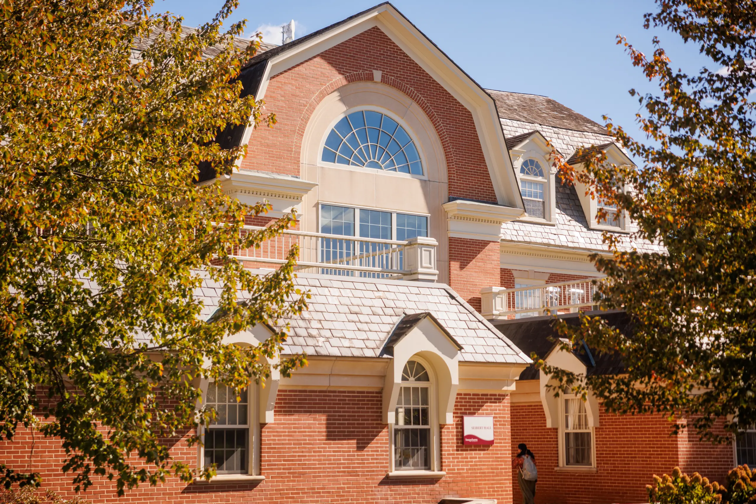 A red-brick building with large arched windows and a gabled roof is partly obscured by trees with green and yellow leaves. A person stands near the entrance, and a sign is visible by the door. The sky is clear and blue.