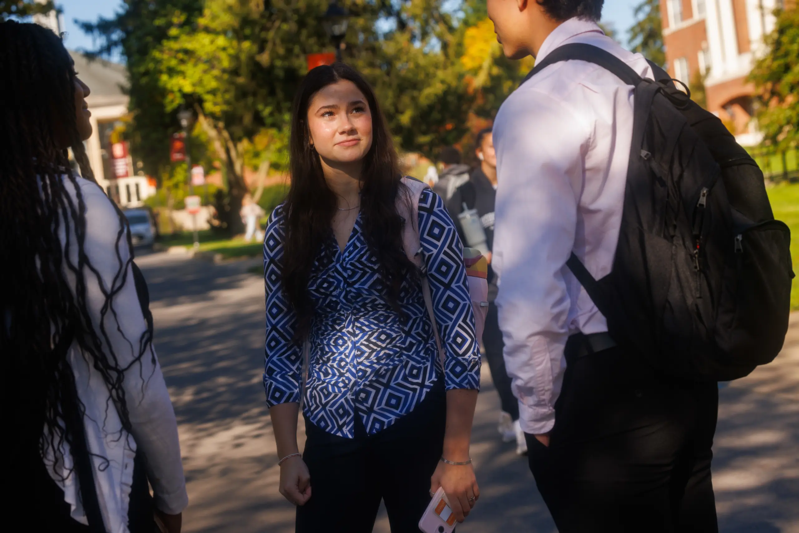A young woman stands outside on a sunny day, talking to two people. She's wearing a blue and white patterned top, holding a phone, and smiling slightly. The background features trees and a building in soft focus.