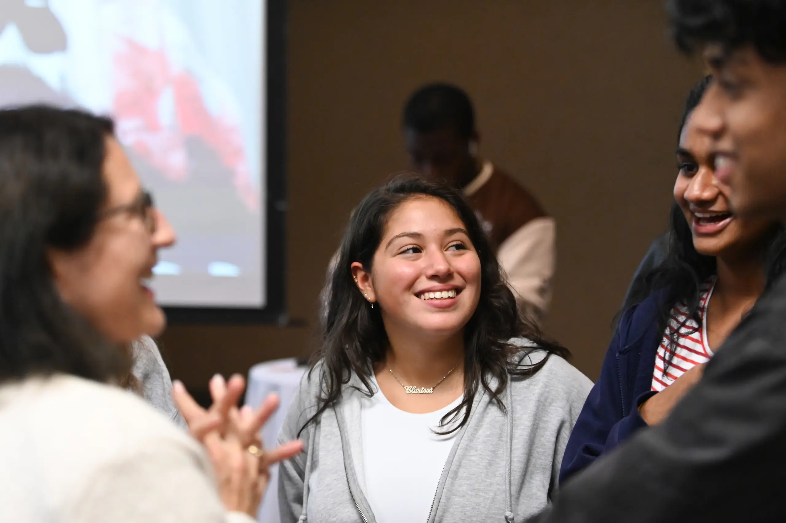 A group of people are gathered indoors, smiling and chatting. One person in the center, wearing a gray sweater and a necklace, is looking to the left. A blurred screen is visible in the background.