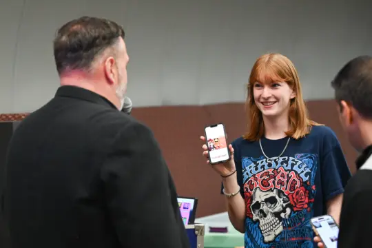 A woman with red hair smiles, holding up a smartphone displaying an image. She's wearing a black T-shirt with a colorful graphic design. A man with a microphone, seen from the back, faces her. Another person stands nearby.