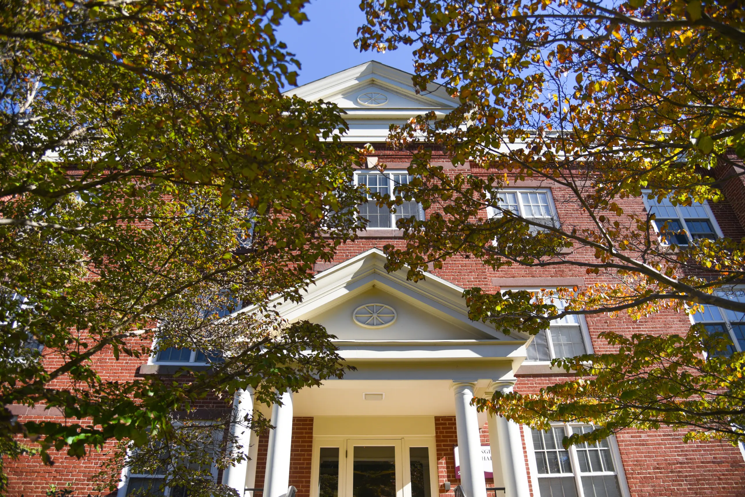 Red brick building with white columns and triangular pediments, partially obscured by green and yellow leaves from surrounding trees. Clear blue sky in the background.