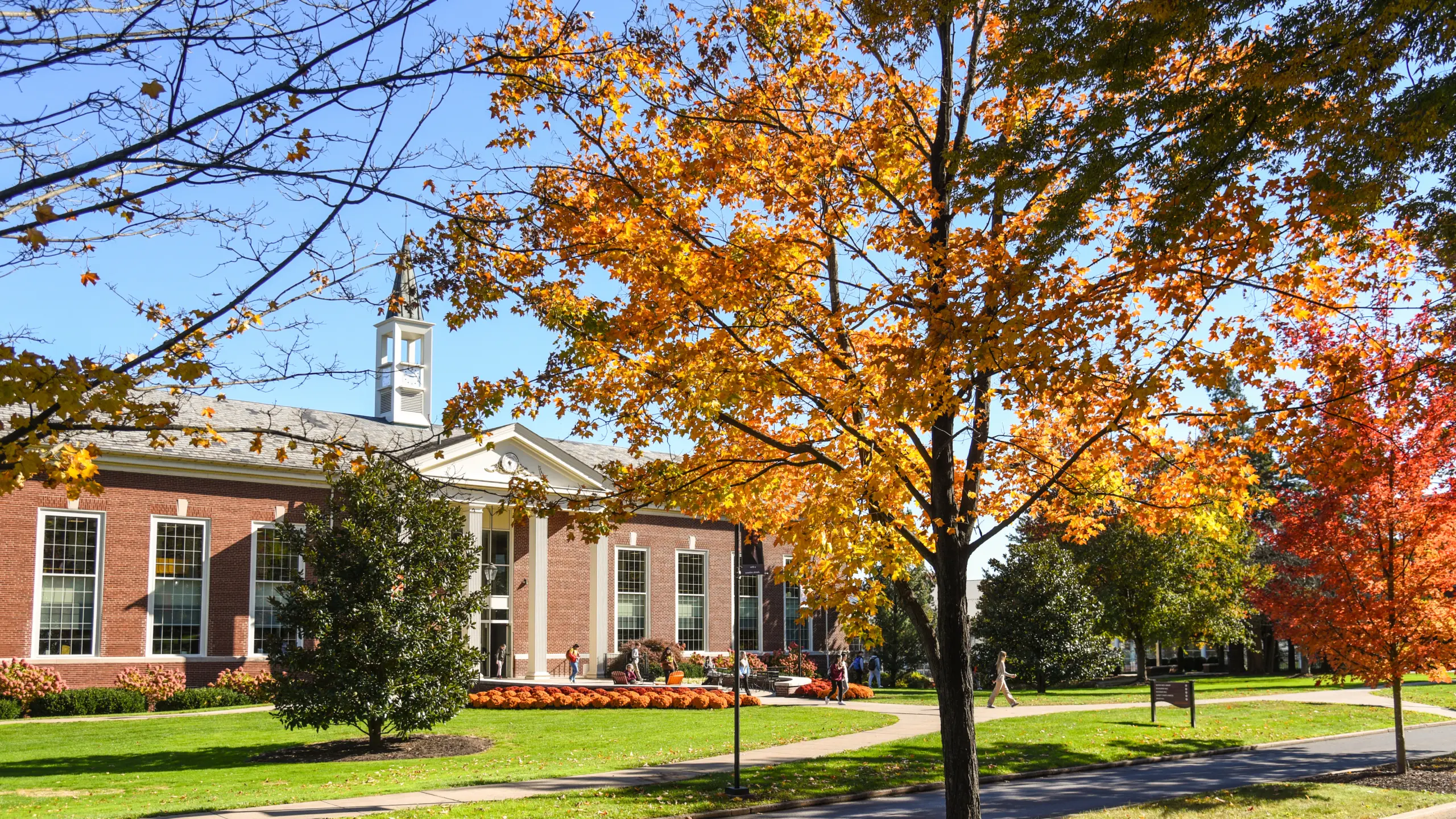 A brick building with large windows and a small spire, surrounded by autumn trees with vibrant orange and yellow leaves. A row of pumpkins decorates the entrance, and the grass is lush and green under a clear blue sky.