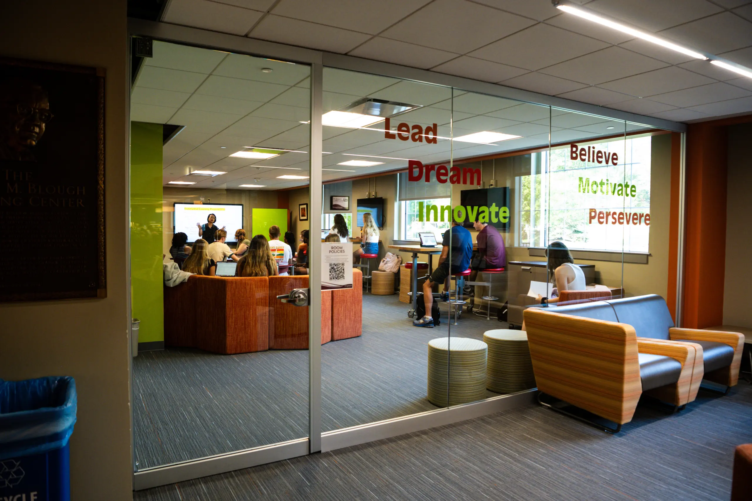 Students are gathered in a modern, glass-walled room with motivational words like 