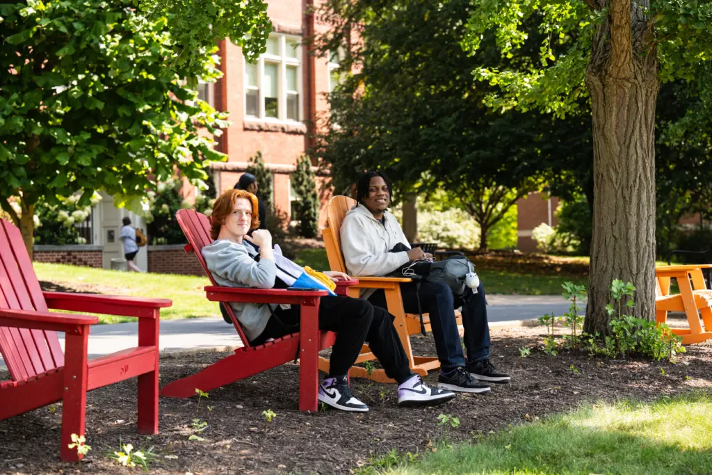 Two people sit on red and orange Adirondack chairs under a tree on a sunny day. One holds a notebook. In the background, there's a brick building with large windows and lush green trees.