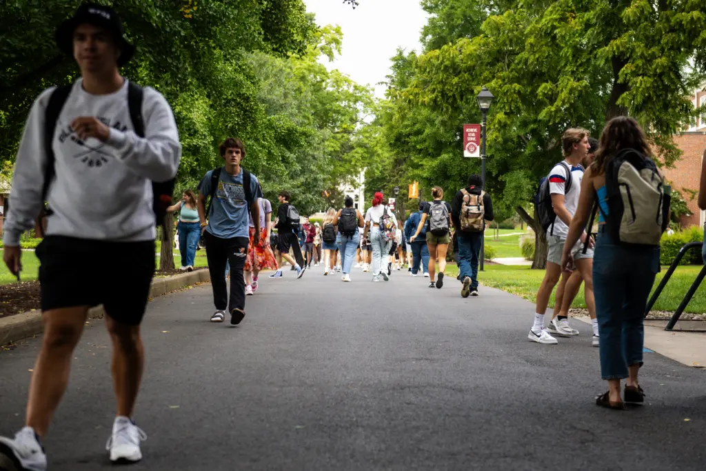 A group of young adults walk along a tree-lined path on a college campus. They carry backpacks and wear casual summer clothing. The background features greenery and a campus banner. It's a bright, sunny day.