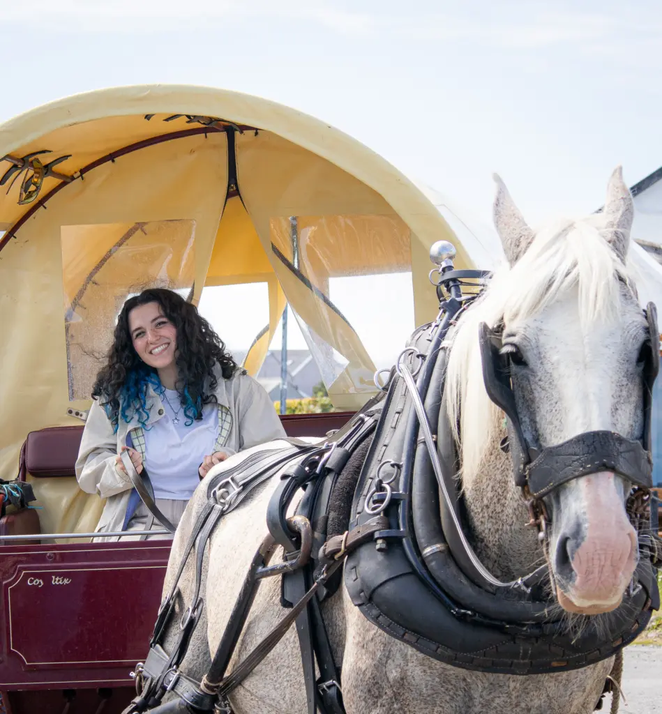 A woman with curly hair smiles while standing next to a gray horse. The horse is hitched to a yellow-covered wagon. The scene suggests a rural or outdoor setting, with a clear sky in the background.