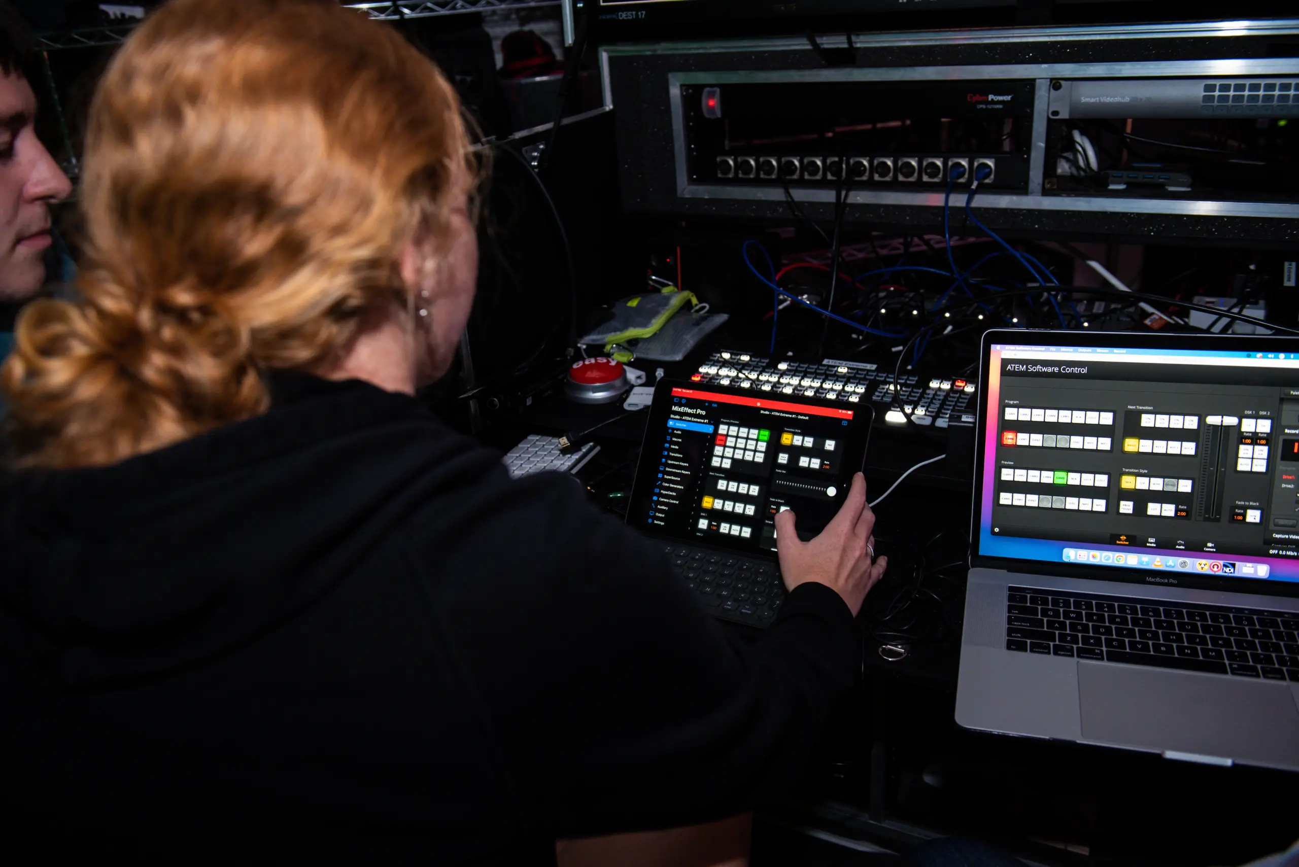 A person with red hair operates a tablet showing various controls, alongside a laptop with a similar interface. Both devices are linked to a complex setup of audio-visual equipment and cables in a dimly lit environment.