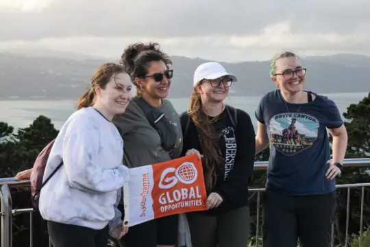 Four students stand together outdoors, smiling and holding a sign reading 