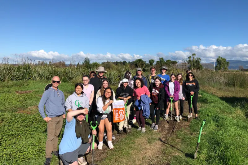 A group of people stand together outdoors in a grassy area under a blue sky. They are holding a 