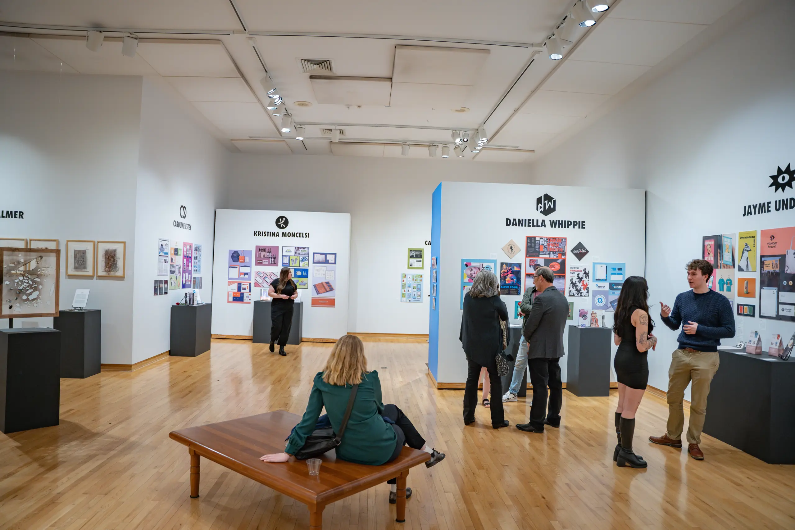 People are viewing art in a modern gallery with light wood flooring. Various artworks are displayed on white walls and black stands. Some individuals are seated on a wooden bench, while others are standing and engaging in conversation.