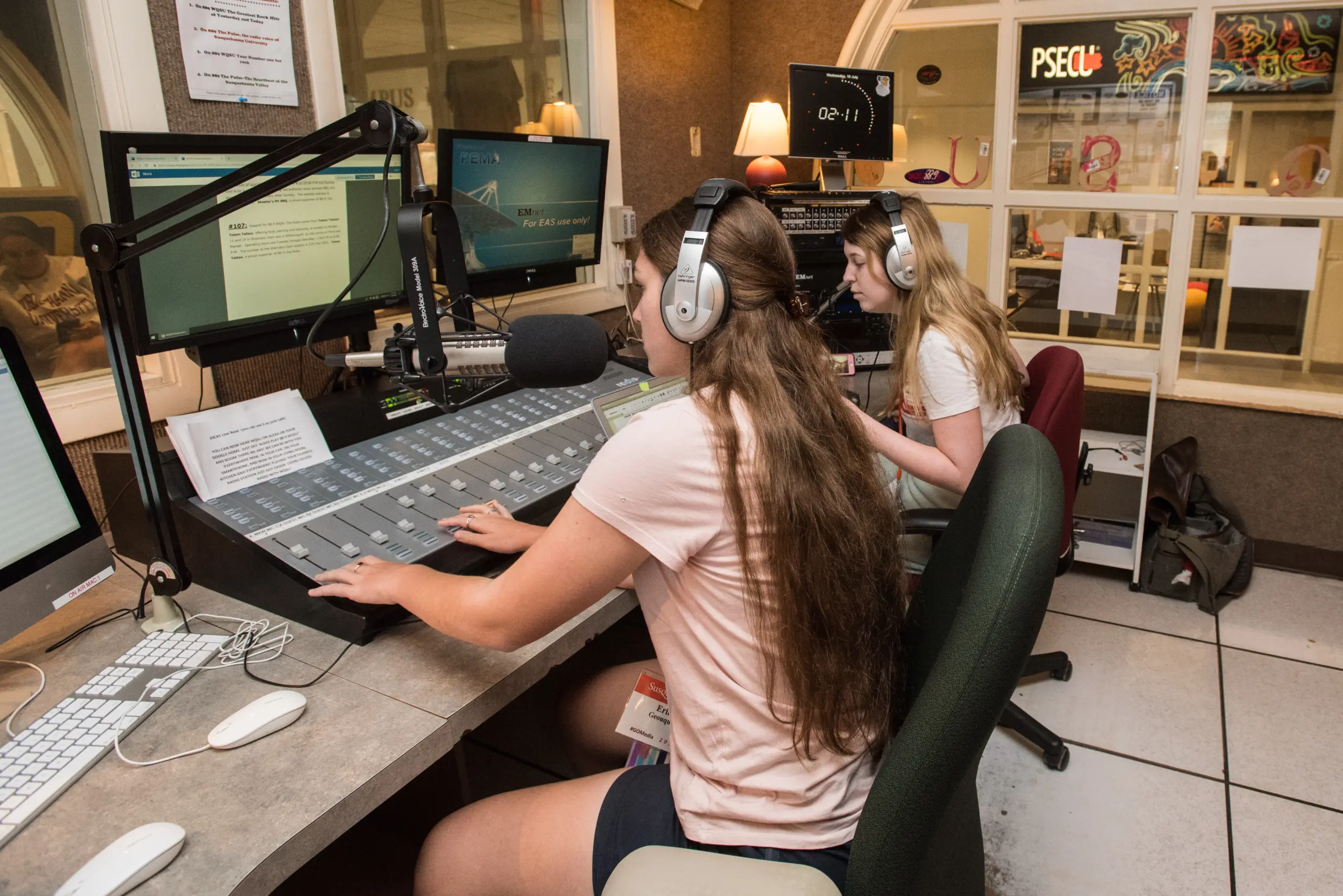 Two people wearing headphones sit at a radio station control board. The person in the foreground has long hair and operates the mixing console. The person in the background is focused on a computer screen. Studio equipment surrounds them.