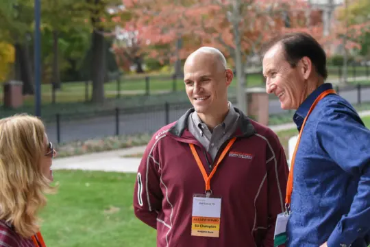 Three people are outdoors on a grassy area with autumn trees in the background. Two men, wearing conference name tags and lanyards, are conversing with a woman wearing glasses. They appear to be enjoying a light-hearted discussion.