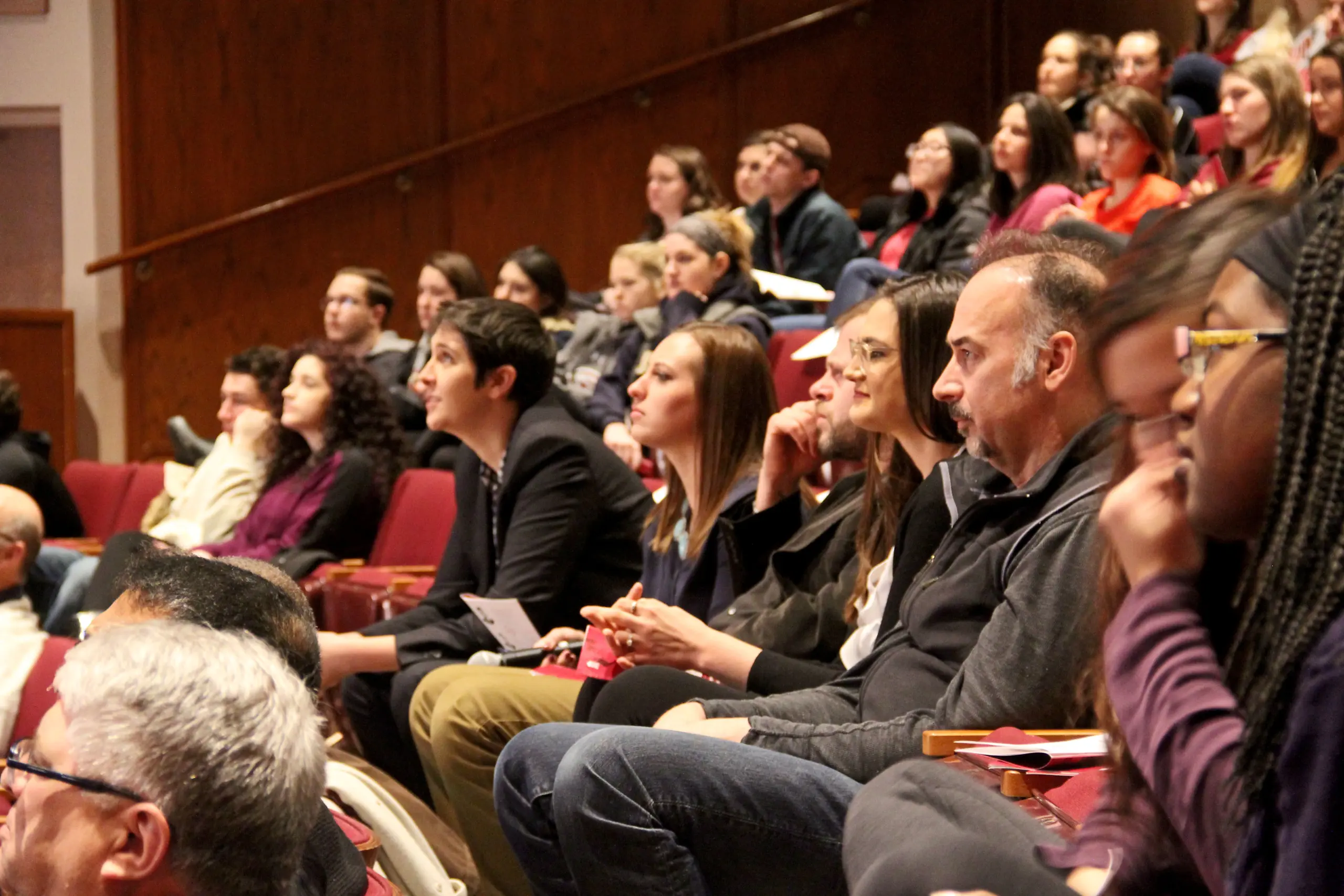 A diverse audience attentively listens during a lecture or presentation in an auditorium. People are seated in tiered rows, with some holding notes. The setting is warmly lit, emphasizing engagement and interest.