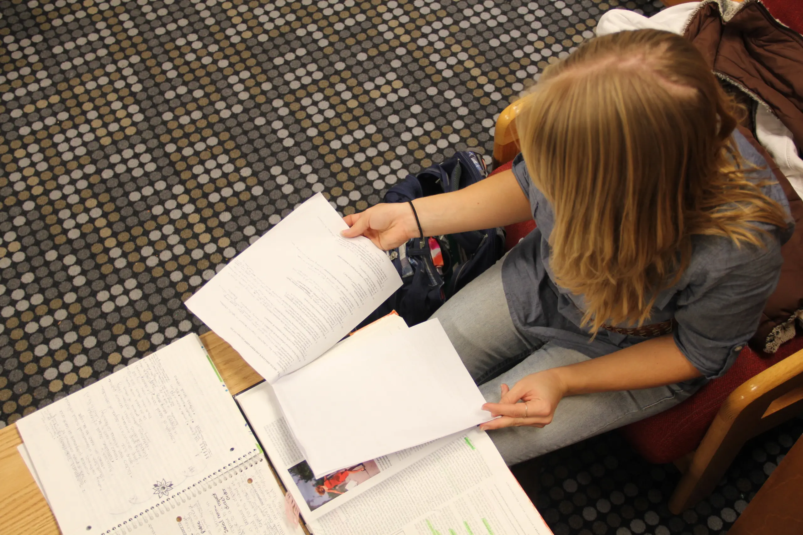 A person with long hair sits in a chair, studying. They are holding papers and looking at open notebooks and a textbook on a table. The floor has a patterned carpet of dark and light dots.