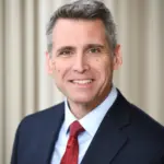 A man with short gray hair, wearing a dark suit, light blue shirt, and red tie, smiles at the camera. The background is out of focus with beige vertical lines.