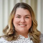 A person with shoulder-length wavy hair smiles at the camera. They are wearing a white blouse with black polka dots and drop earrings. The background is softly blurred with vertical lines.
