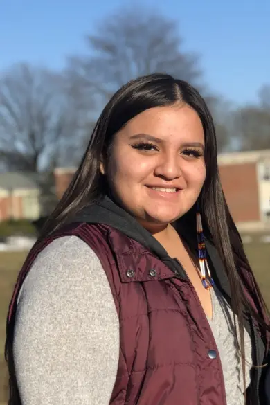 A woman with long brown hair, wearing a maroon vest and gray top, smiles while standing outdoors. A tree and a building are visible in the background under a clear blue sky.