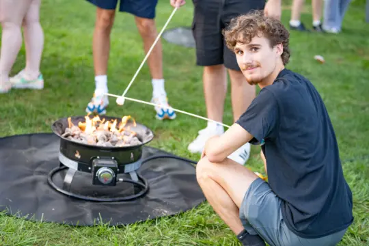 A young man crouches by a portable fire pit on a grassy area, holding a marshmallow on a stick over the flames. Other people, partially visible, are also roasting marshmallows. Everyone is casually dressed, enjoying an outdoor gathering.
