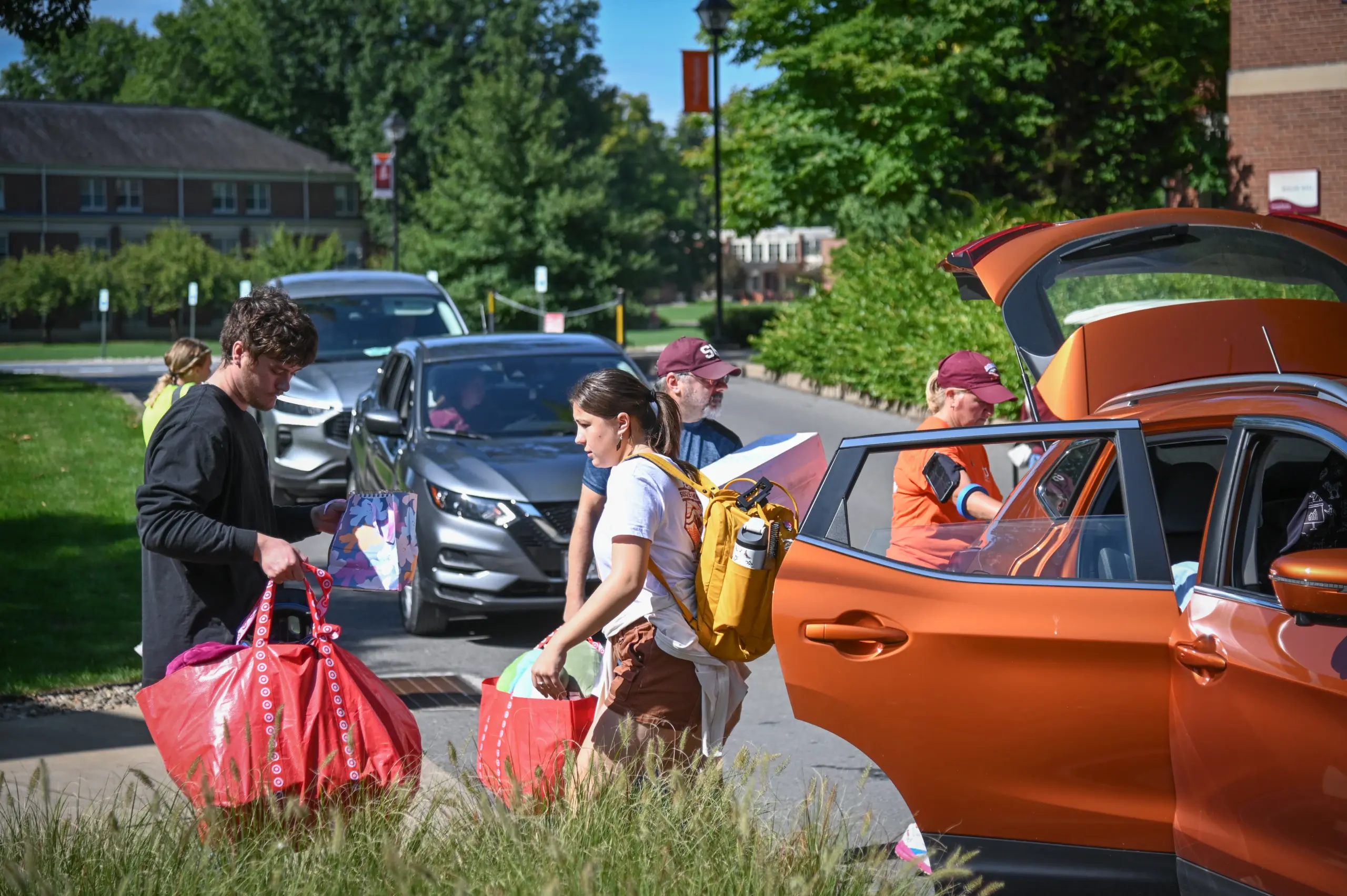 People unload bags and items from an orange car outside a university dorm. A young woman carries a backpack, and others assist with the luggage. Cars are parked along the street, and trees and a building are visible in the background.