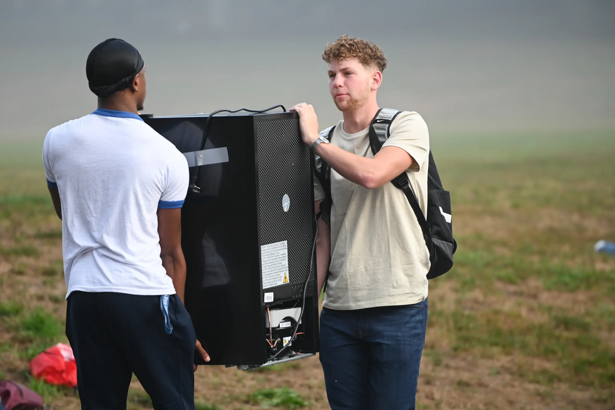 Two men are outdoors carrying a large, black electronic device with handles on the side. One man wears a white shirt and cap, the other a beige shirt and backpack. They stand on a grassy field with a misty backdrop.