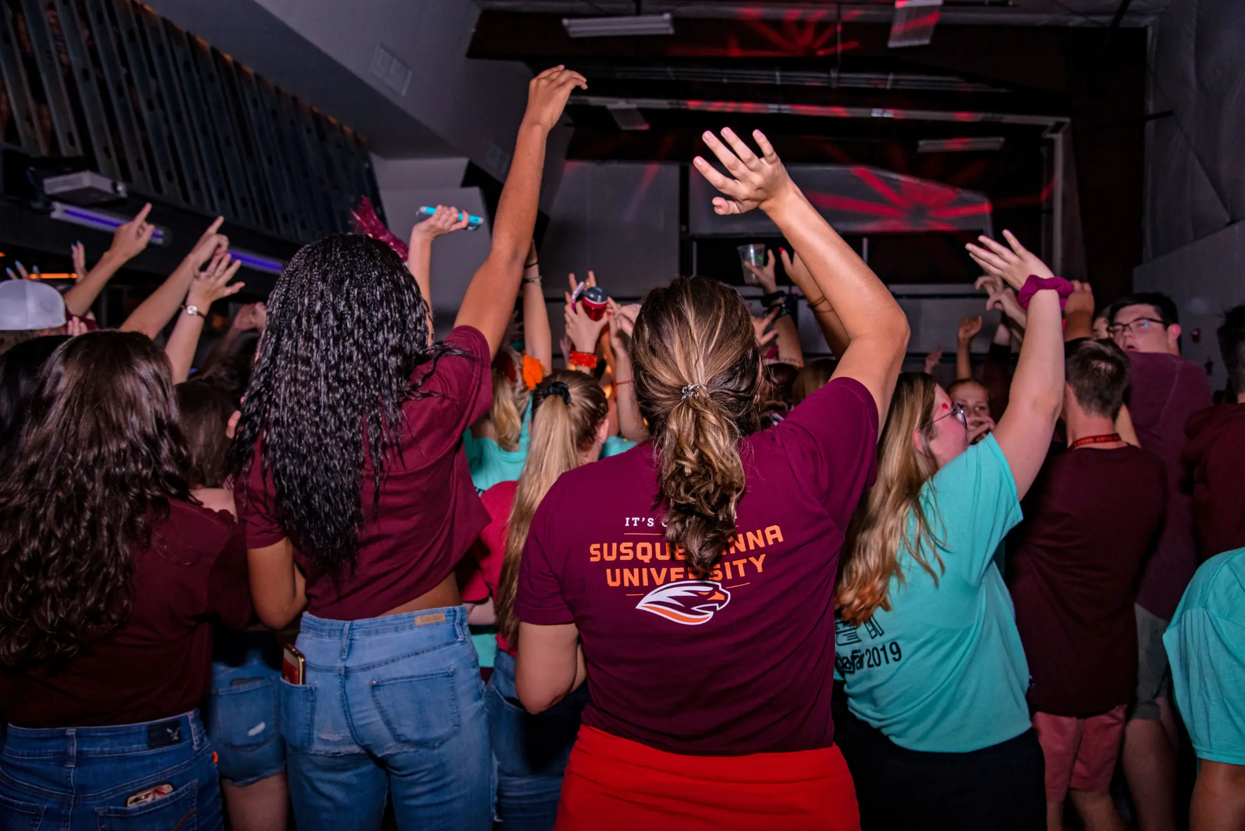 A lively crowd of people in casual wear dance and raise their hands at an indoor event. Several individuals are wearing maroon and turquoise shirts, with one shirt displaying 