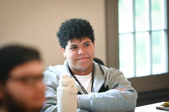 A person with curly hair, wearing a gray hoodie, sits with arms crossed at a table. They appear thoughtful and are in a room with a blurred figure in the foreground and a window in the background.