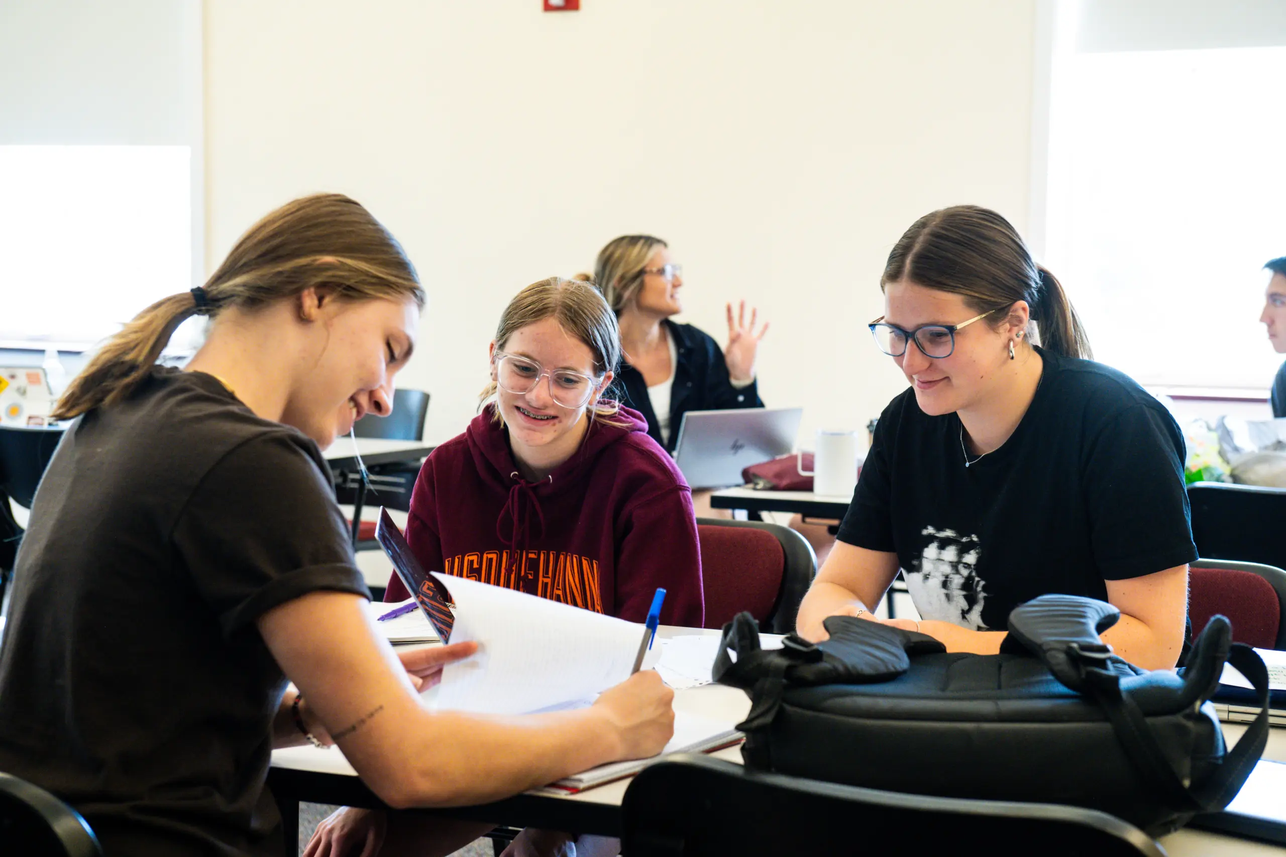 Three students are sitting together in a classroom, working on an assignment. They are smiling and appear engaged. One student is writing in a notebook while the others look on. More students are visible in the background.