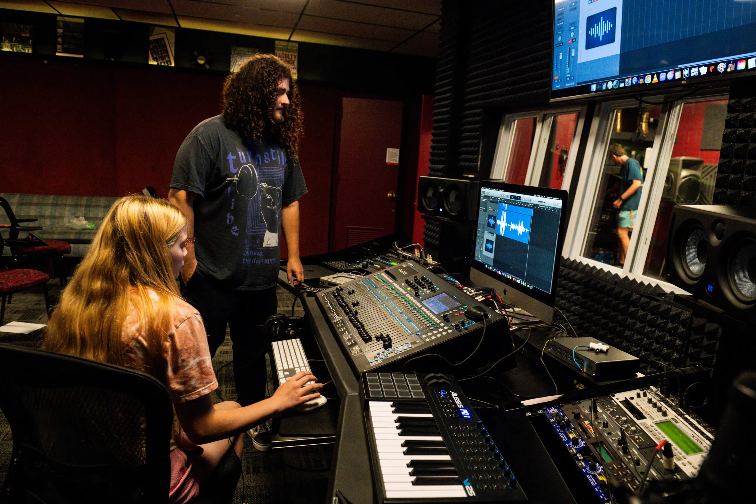 A woman sits at a control board operating audio equipment in a recording studio, while a man stands beside her observing. Monitors and various sound equipment are visible in the dimly lit room.