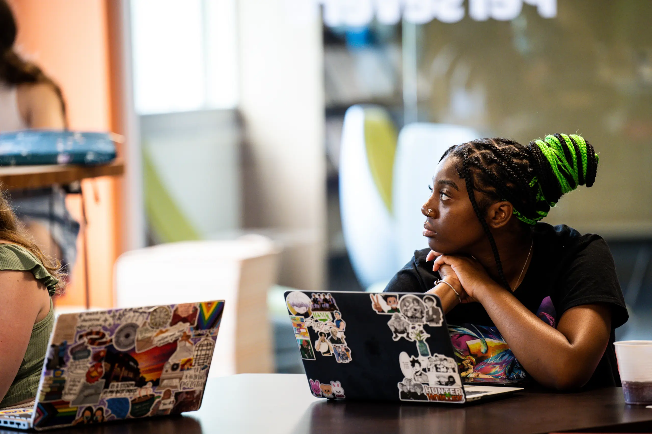 A person with green braided hair looks to the side, possibly in thought, during a lively Writers Workshop. They sit at a table with a laptop covered in various stickers. Another participant with a similar laptop is partially visible to the left. The room is bright and modern, fostering creativity.