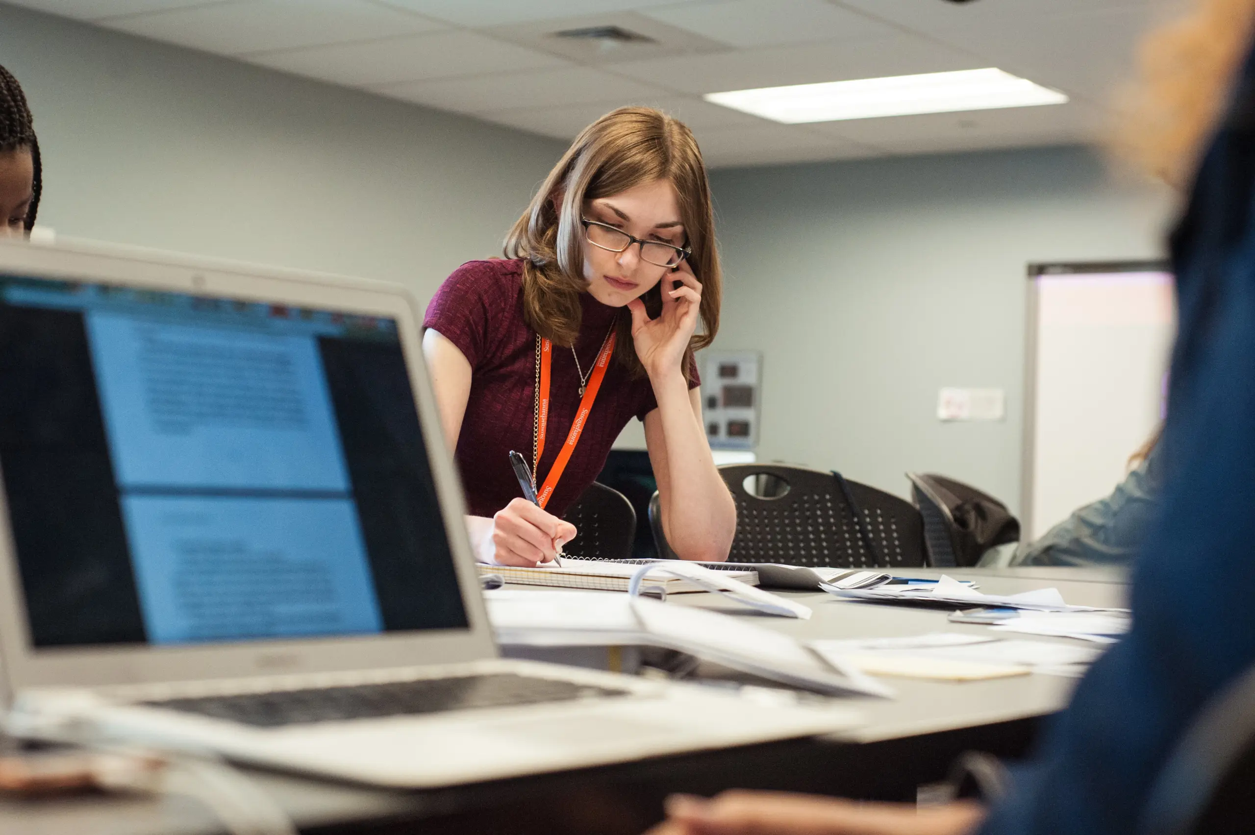 A young person with glasses and a red lanyard sits at a desk, engrossed in writing notes in a notebook. Holding a phone to their ear, they channel the focus of The Quill. A laptop with an open document sits nearby, while chairs linger in the background.