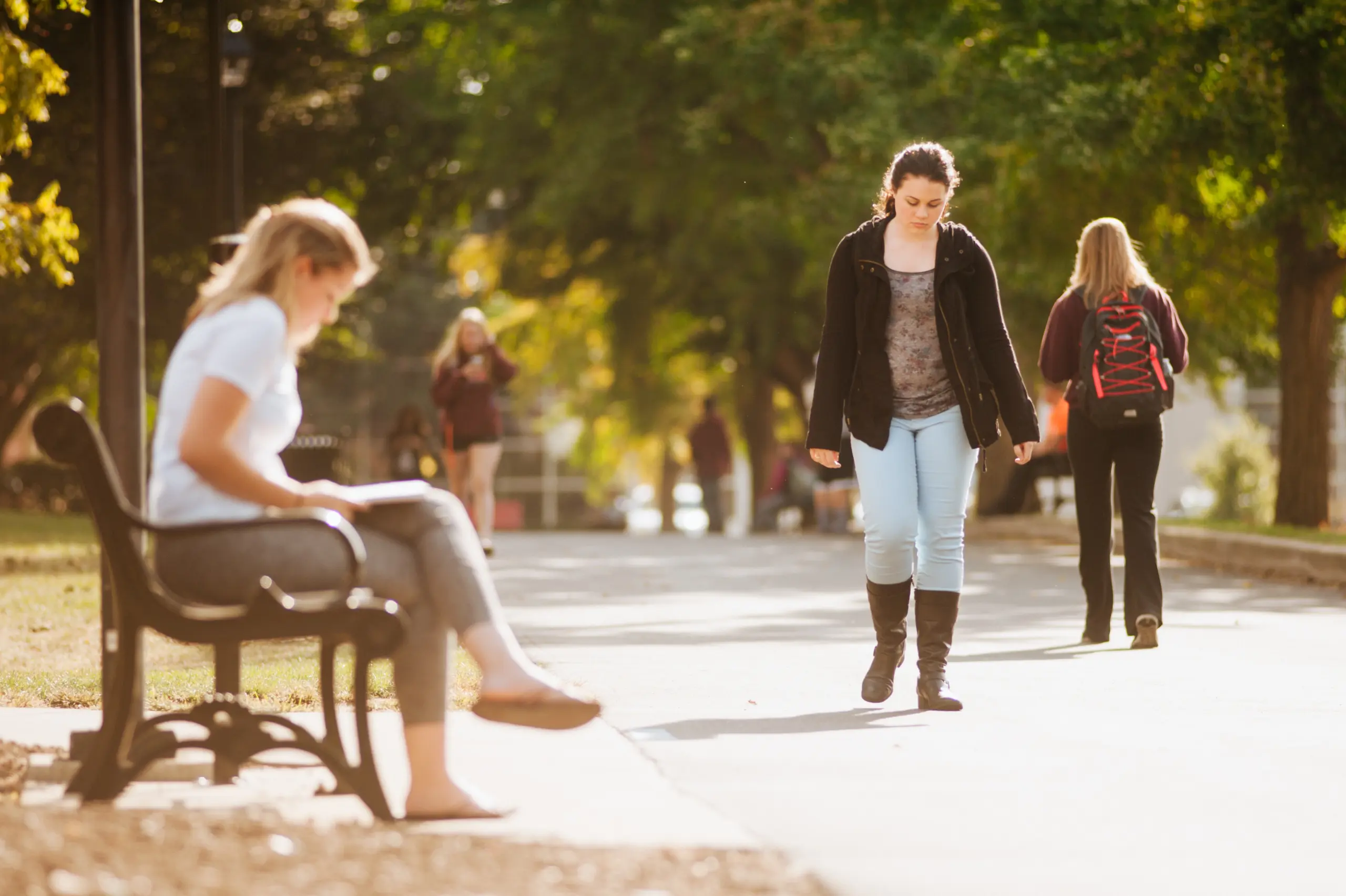 A woman walks down a sunlit path lined with trees. Another woman sits on a bench in the foreground, reading. Other people stroll in the background, enjoying a sunny day in a park-like setting.