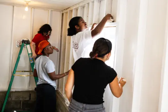 Four people painting a white wall inside a room. One person is on a ladder painting near the ceiling, while the others work on different areas of the wall. The room has a bare light bulb hanging from the ceiling.