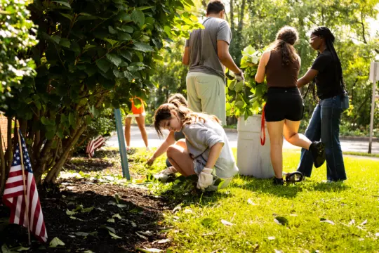 A group of people gardening on a sunny day, surrounded by lush greenery. One person kneels to tend to plants, while others stand, holding leaves and bags. Two small U.S. flags are visible in the foreground.