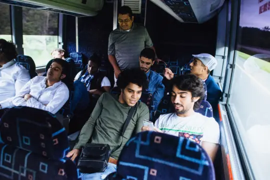 A group of people sitting on a bus, some looking at a smartphone, while others are relaxing and chatting. The seats are patterned, and outside the window, greenery is visible.