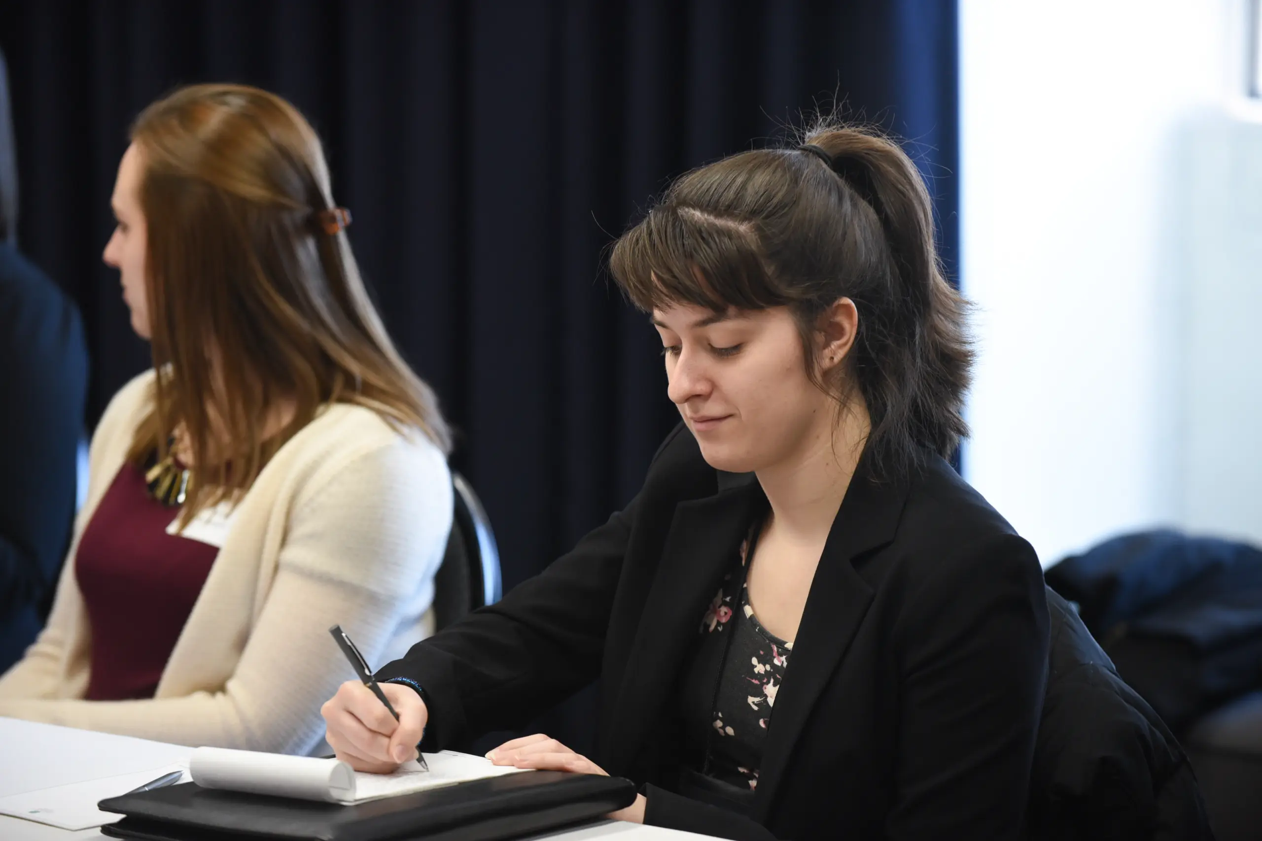 A woman with dark hair in a ponytail, wearing a black blazer, diligently writes notes at a table. Her fellow apprentice writer, sporting brown hair and a white cardigan, sits beside her. They are in a room with dark curtains that add to the focused atmosphere.