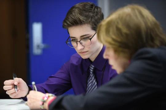 Two individuals are seated and engaged in an activity involving writing or reviewing documents. The person in the foreground wears glasses and a purple shirt, and appears focused. The background is slightly blurred, featuring a door with a blue wall.
