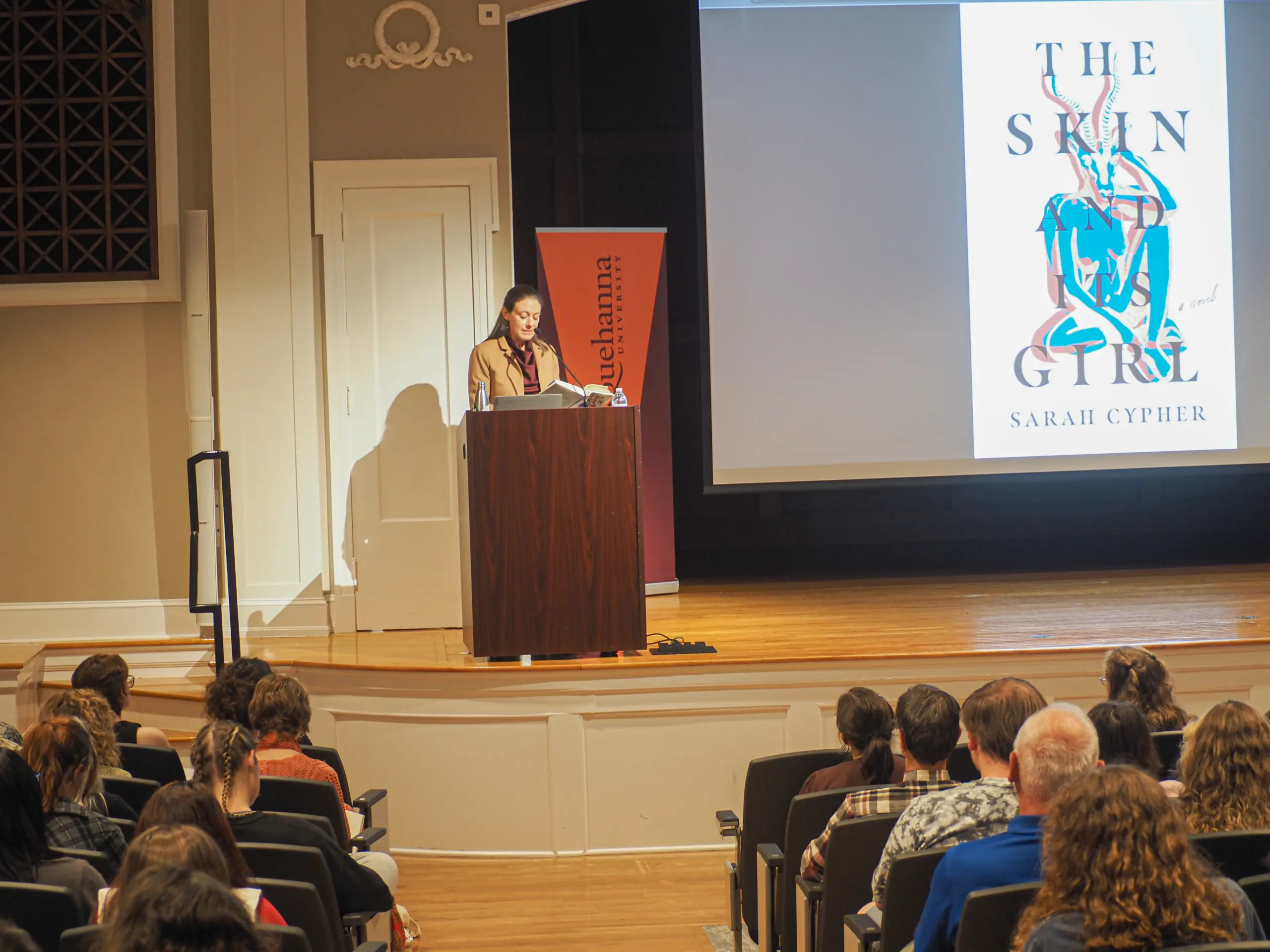 A person stands at a podium giving a lecture or presentation to an audience. On a screen, the book cover 