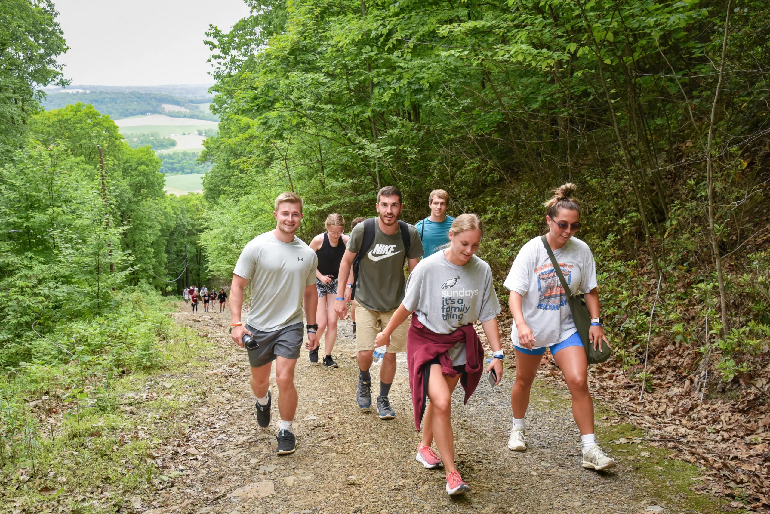 A group of people engaged in outdoor recreation is hiking up a forest trail. They are dressed casually, with some in shorts and t-shirts. Lush green trees line the path, and a scenic landscape unfolds in the background. Smiles abound as they enjoy their invigorating walk.