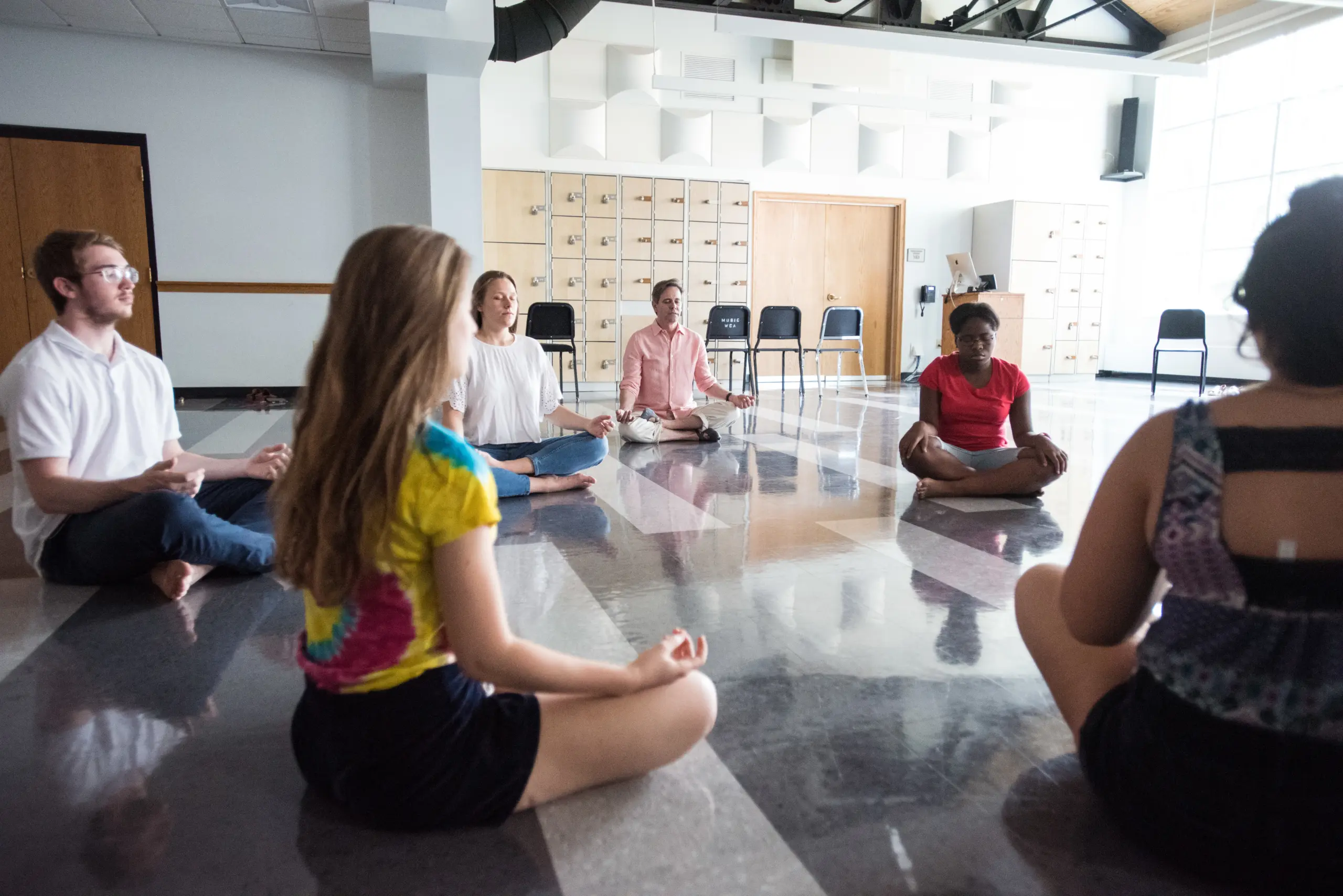 A group of people sitting in a circle on the floor of a spacious room, participating in a meditation, yoga, or mindfulness session. They are seated with legs crossed and eyes closed. Chairs and lockers line the wall in the background.