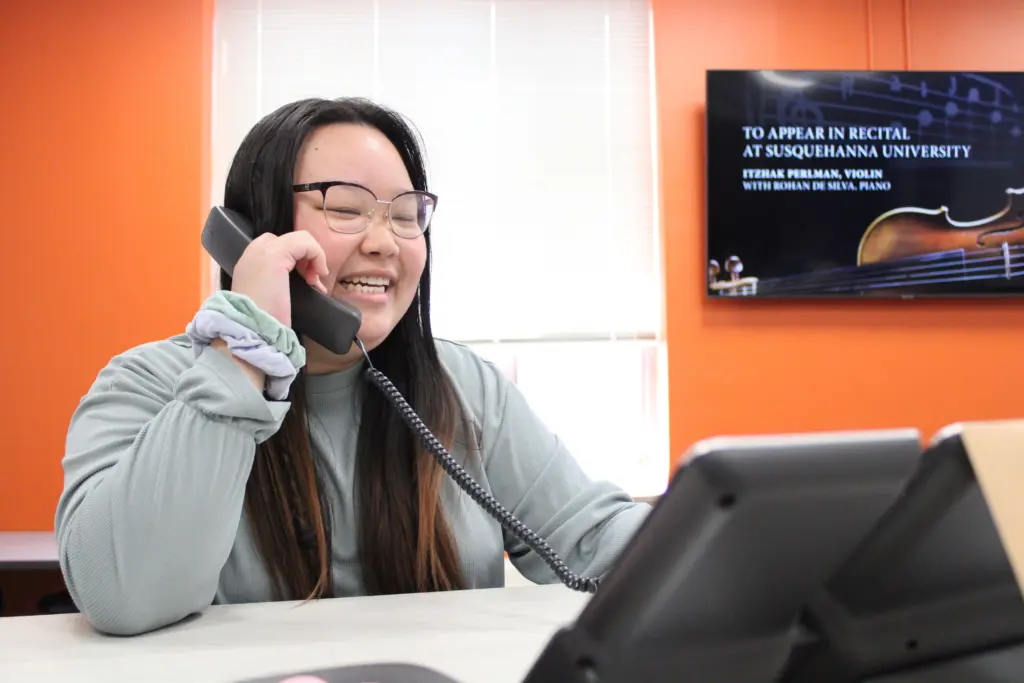 A smiling person with long dark hair and glasses chats on a landline phone in an office with orange walls, likely discussing student employment opportunities. A poster on the wall announces a recital at 体育买球官网 University.