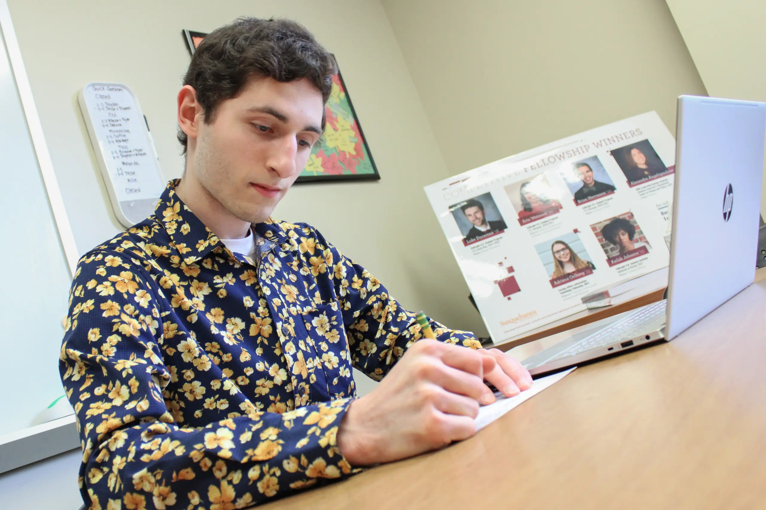 A person in a floral shirt is writing on paper at a desk, their laptop open and displaying student employment opportunities. A poster with multiple faces adorns the background, adding character to the office or study space where this scholarly pursuit unfolds.